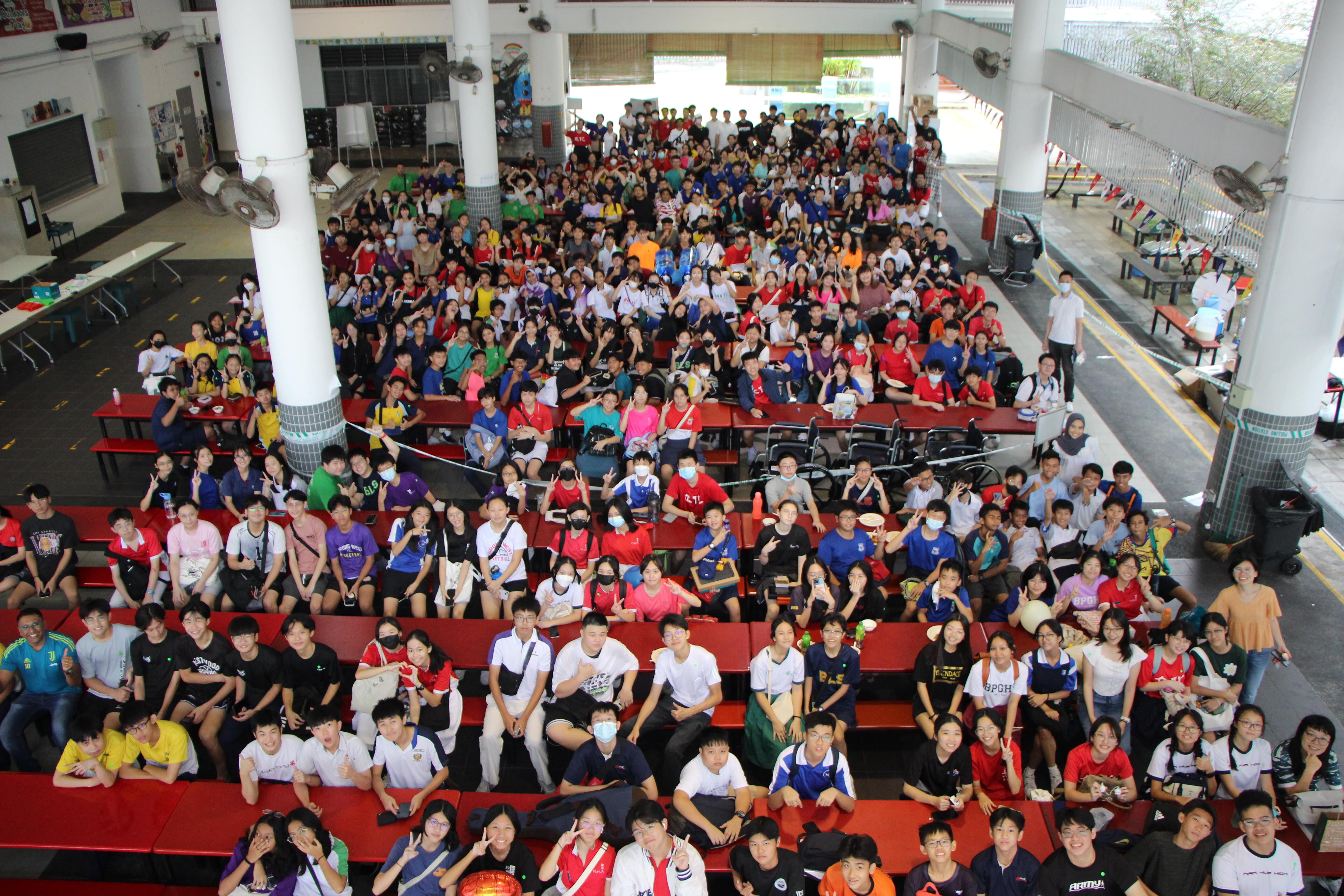 Large group of students in colorful attire seated on red benches in a school canteen, posing for a photo.